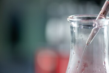 A close up of a wide-mouth graduated conical flask, three-quarters full with a transparent red liquid, sitting on a lab bench with a test tube rack full of similar red liquid-filled test tubes in the