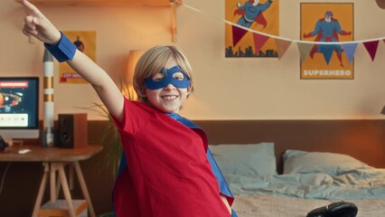 Medium slowmo of joyful Caucasian boy in superhero costume with eye mask and cloak spinning and raising his hand in air while playing at home during leisure time
