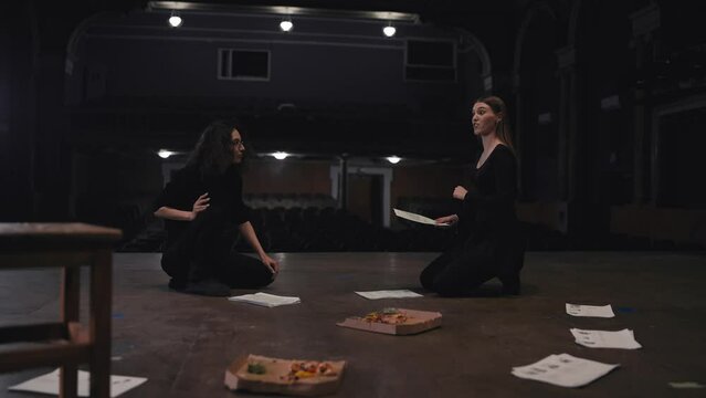 A confident girl with brown hair and a black suit holds sheets of paper with a script in her hands and expressively addresses her fellow actress during her rehearsal for a play on the theater stage