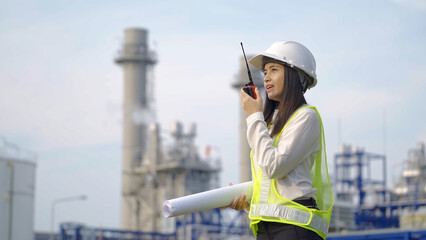 A female engineer in a helmet and reflective vest uses a walkie-talkie while holding blueprints at...
