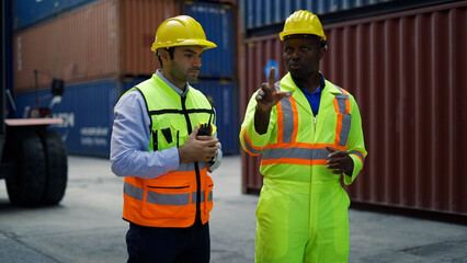 Two workers wearing hard hats and safety vests discuss logistics and operations at a shipping container yard with stacked containers in the background.  