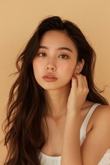 The young Asian beauty's long curly hair with Korean makeup style touches her face and perfect skin against an isolated beige background. Facial care, beauty, plastic surgery.