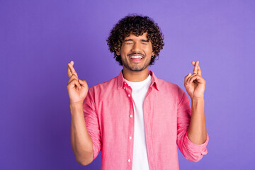 Portrait of nice young man crossed fingers closed eyes wear pink shirt isolated on purple color...