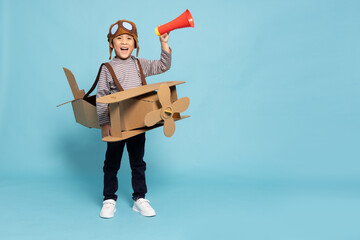 Asian boy aviator holding megaphone and playing with cardboard airplane isolated on blue sky background, Announce speech by kids with toy concept, Six years old