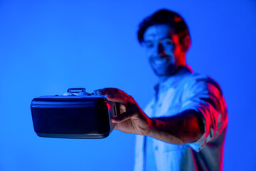Smiling caucasian man holding and show VR headset with blurring background while standing with neon...