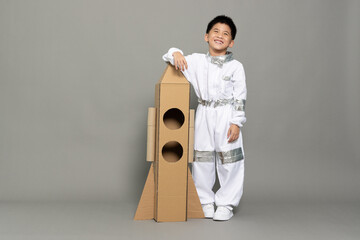 Asian little boy pilot or spaceman smiling and standing with cardboard rocket isolated on grey...