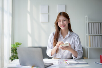 Smiling woman working from home, holding notepad, and sitting at a desk with laptop in a bright...