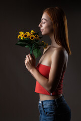 A peaceful portrait of a young woman enjoying the scent of sunflowers, dressed in a stylish red top against a muted background.