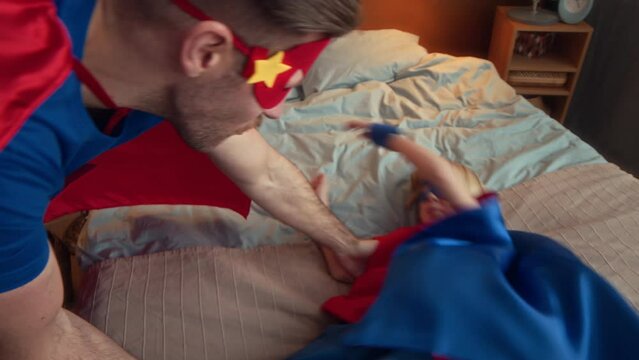 Tracking slowmo of playful Caucasian tween boy and his father both wearing cute red and blue superhero suits having fun at home together, jumping off to bed and laughing