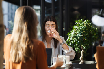 Two young business women in a cafe having one on one meeting. Friends after work talking gossiping and having coffee at a window table with reflections..