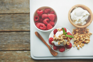 Yogurt, bowl and glass with raspberry in cereal for breakfast food, nutrition and snack in morning. Top view, fruit and dairy in container with wooden spoon for healthy meal, organic and vegan diet
