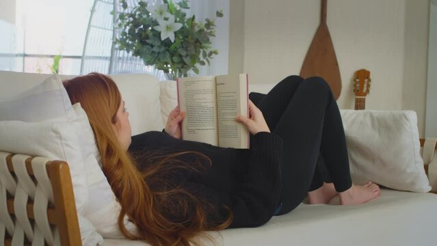 Cinematic video of a young woman reading a book and relaxing on a sofa in the living room. Red-haired girl lying down to read a storybook. Video concept of woman enjoying her free time, resting.