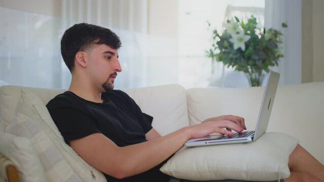 Relaxed serious millennial guy student freelancer using laptop on couch at home. Focused young man working distantly typing on notebook in apartment.