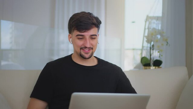 Happy young man putting earphones talking video calling on chatting laughing enjoy online conversation looking at webcam. Millennial guy working from home having a work meeting.