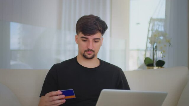 Young man shopping online with a credit card. Millennial guy makes payment via on-line web store using laptop. Man spending money on internet, e-shopping at home.