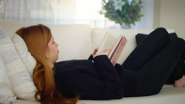 Young Woman Lying on Couch Reading a Book. Girl with red hair is lying on a white couch, engrossed in reading a book, enjoying a peaceful moment