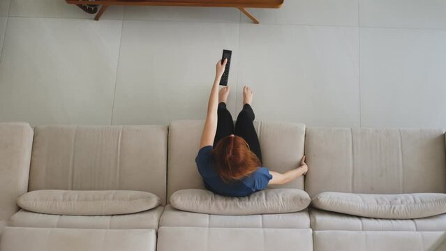 Top View of Woman Relaxing on Couch and Watching TV. Woman with red hair sits on a couch, grabs the remote control, and turns on the TV, then lies down to enjoy her free time. Leisure and relaxation