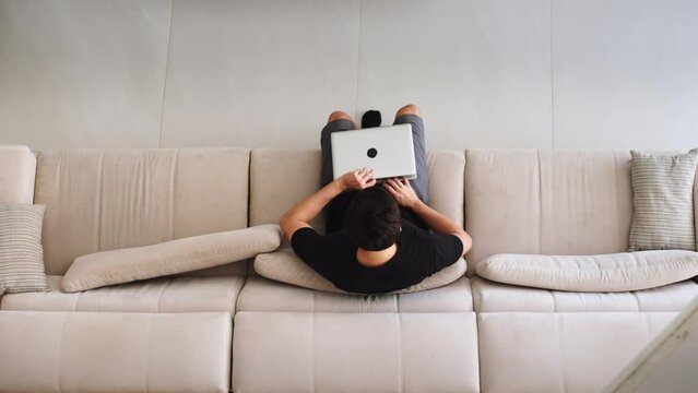 Top View of a Man Working on a Laptop on a Sofa. Man Sitting on a Couch Using Laptop in a Modern Home Setting. Student studying online through laptop at home.