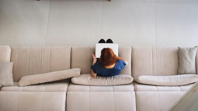 Woman Working on a Laptop on a Sofa. Girl Sitting on a Couch Using Laptop in a Modern Home Setting. Student studying online through laptop at home