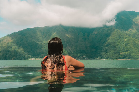 A young woman on vacation relaxing in the summer at hot thermal springs in a mountainous area. She sits in the pool and looks into the distance at the hills and lake that is located in front of her.