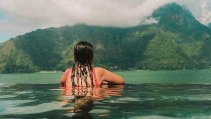 A young Caucasian brunette woman in a red swimsuit on vacation relaxes in a resort with an infinity pool overlooking the mountains and lake. Summer holidays and wellness treatments for the body.
