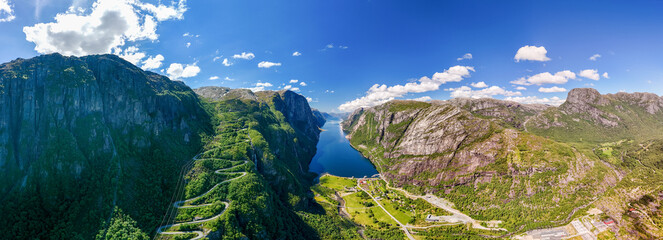 view of a winding road snaking through a stunning Norwegian fjord. Lush green mountains and a deep blue body of water create a breathtaking landscape under a clear blue sky. Lysefjord, Norway - Powered by Adobe
