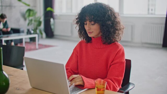 Side view of Caucasian female with dark curly hair sitting at desk. Working on laptop. Typing something on keyboard. Pressing keys. Working remotely. Wearing warm red sweater. Office or apartment.