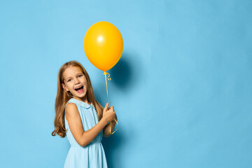 Fototapeta premium Joyful little girl in a summer dress holding a yellow balloon against a vibrant blue background