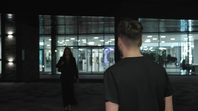 Guy meets his girlfriend in the evening after work and kisses her. The businesswoman has finished her work and goes outside at night. Medium shot.