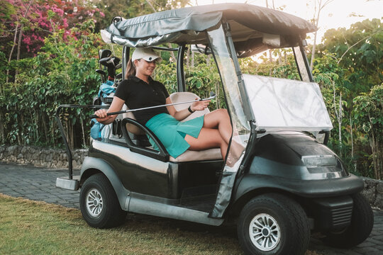 Young attractive professional female golfer sitting in a golf cart next to the green wearing sportswear and a cap. She smiles and looks at her golf club. Preparing for competitions at the country club
