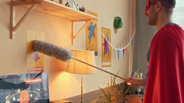Medium side view shot of Caucasian young adult man dressed up in red and blue superhero costume cleaning shelves with pp duster, doing housework in child bedroom