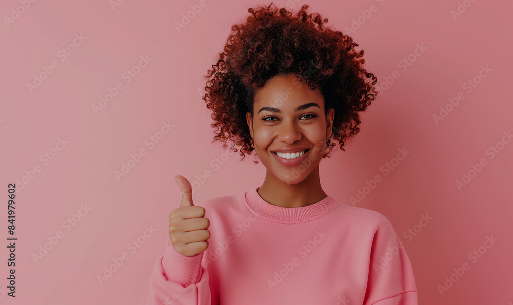 Wall mural Young curly african american girl in a pink sweatshirt smiling with healthy teeth showing thumb up at copy space expressing wow emotion standing isolated on pink background - Wall murals
