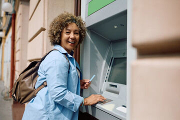 Obraz premium Happy female tourist entering pin while using ATM machine and looking at camera.