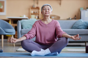 Full length portrait of grey haired senior woman enjoying yoga practice at home and meditating with eyes closed in lotus position copy space