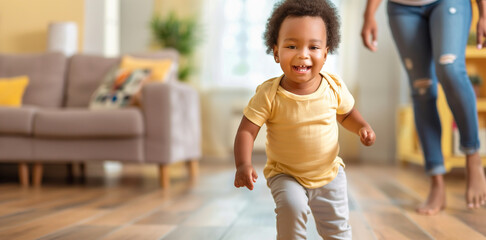 Cute toddler taking his first steps in bright and sunny living room, with hist parent watching in awe in background.