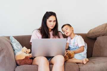 Young Mother and son on the sofa using laptop. Happy mom and little boy hugging and watching videos, reading books, playing video game, using online app, studying together at home. Family time