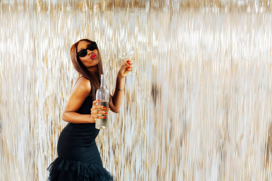 Young happy fashion African girl black woman in cocktail dress and sunglasses holding drink glass and wine bottle dancing at party with glitter background having fun at holiday, birthday celebration.