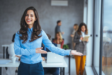 Obraz premium A smiling Caucasian businesswoman stands confidently in an office, arms outstretched in a welcoming gesture, dressed in professional blue attire with colleagues in conversation behind her.