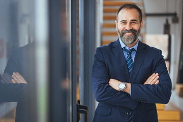 A mature male executive with a beard stands arms-crossed in a well-lit office, dressed in a professional navy suit and tie, exuding confidence and professionalism.