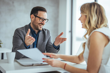 A male and female in formal attire engage in a productive dialogue, sharing documents across the...