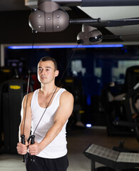 Sportive young man doing arm exercises on adjustable cable training device in well-equipped gym