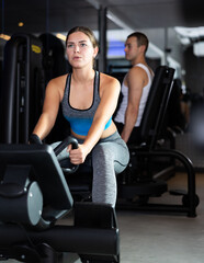 Sportive young woman doing arm exercises on cable training device in well-equipped gym