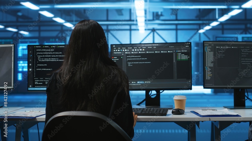 Poster it support employee in data center putting headphones on to talk with coworkers, coding on computer.