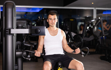 Sportive young man doing exercises on arm curl device in well-equipped gym