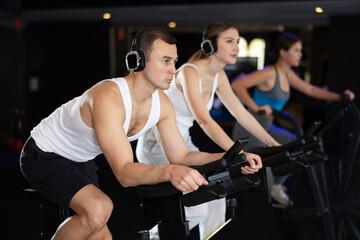 Determined young guy training with spin bike in fitness hall during full-body workout