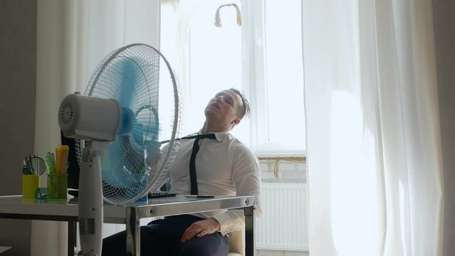 A person in a working office is very hot, a fan cools the person, the sun is shining brightly on the office worker