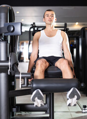Determined young guy training his legs using exercise machine in fitness hall