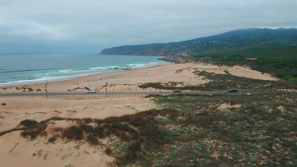 Foamy waves washing beach lined with sandy dunes drone view. Road stretching 