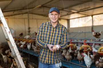 Happy successful farmer engaged in breeding of laying hens and producing organic eggs, posing in...