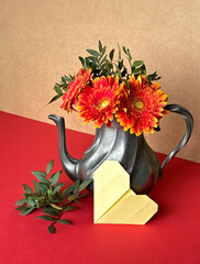Vintage Silver Teapot with Vibrant Orange Gerbera Flowers and Origami Heart on Red Background.